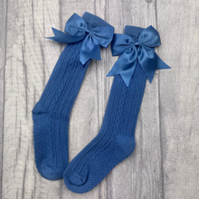 Load image into Gallery viewer, A pair of blue cable-knit socks with matching satin bows at the top, displayed on a gray wood-plank surface.
