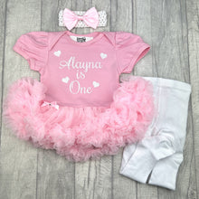 Load image into Gallery viewer, Baby girls light pink tutu romper with bow headband above and white tights on the right side. The tutu romper features white glitter love hearts design and script lettering saying Name is One.
