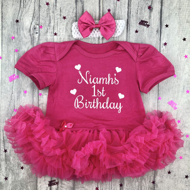 Baby girls dark pink tutu romper with matching bow headband above. The tutu romper features white glitter love heart design with script lettering saying Name 1st Birthday