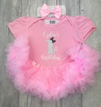 Load image into Gallery viewer, A pink baby girl's first birthday outfit set featuring a short-sleeve bodysuit with 'Lydia's 1st Birthday' text and a glittery number 1, styled with a matching pink tutu skirt and a pink bow headband, laid flat on a gray wood background.
