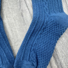 Load image into Gallery viewer, A pair of blue cable-knit crew socks laid flat on a gray wood-textured surface.
