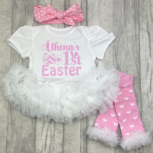 Baby girls short sleeve white tutu romper with pink headband above and leg warmers below. The tutu romper features pink glitter easter egg and bunny ears design, with lettering saying Name's 1st Easter.