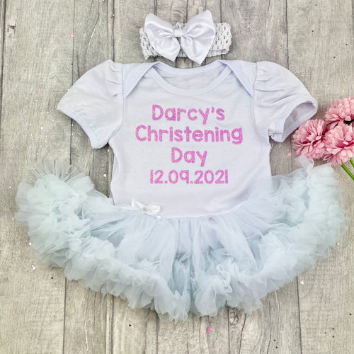 Baby girls short sleeve white tutu romper with bow headband above. The tutu romper features light pink glitter lettering saying Name Christening Day Date