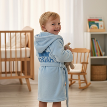 Load image into Gallery viewer, A toddler stands barefoot in a softly lit nursery, wearing a light blue hooded bathrobe with 'OSIAH' printed on the back and holding a plush toy.

